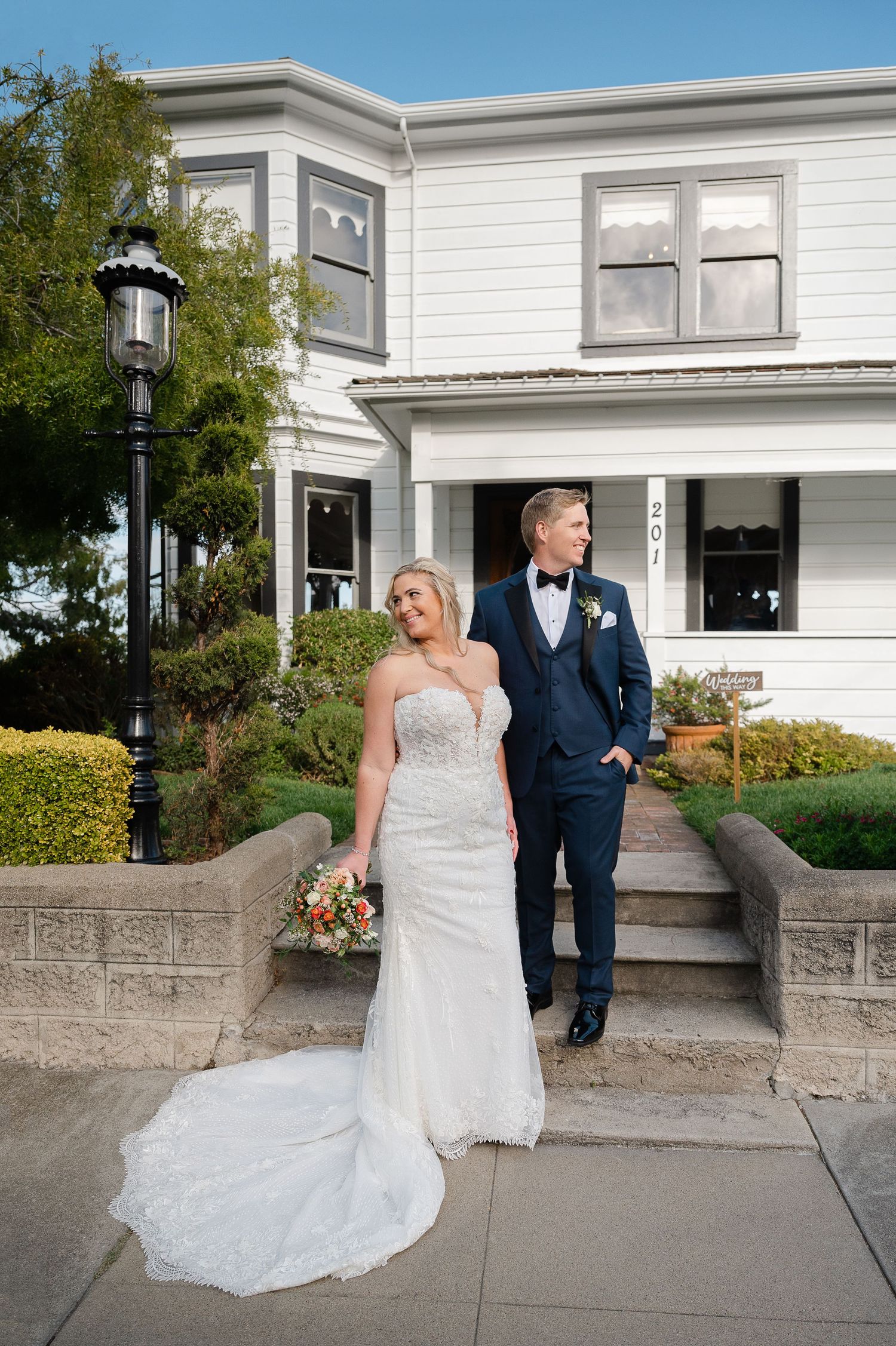 Bride in white gown and groom in navy tuxedo pose in front of a white Victorian house on their wedding day.