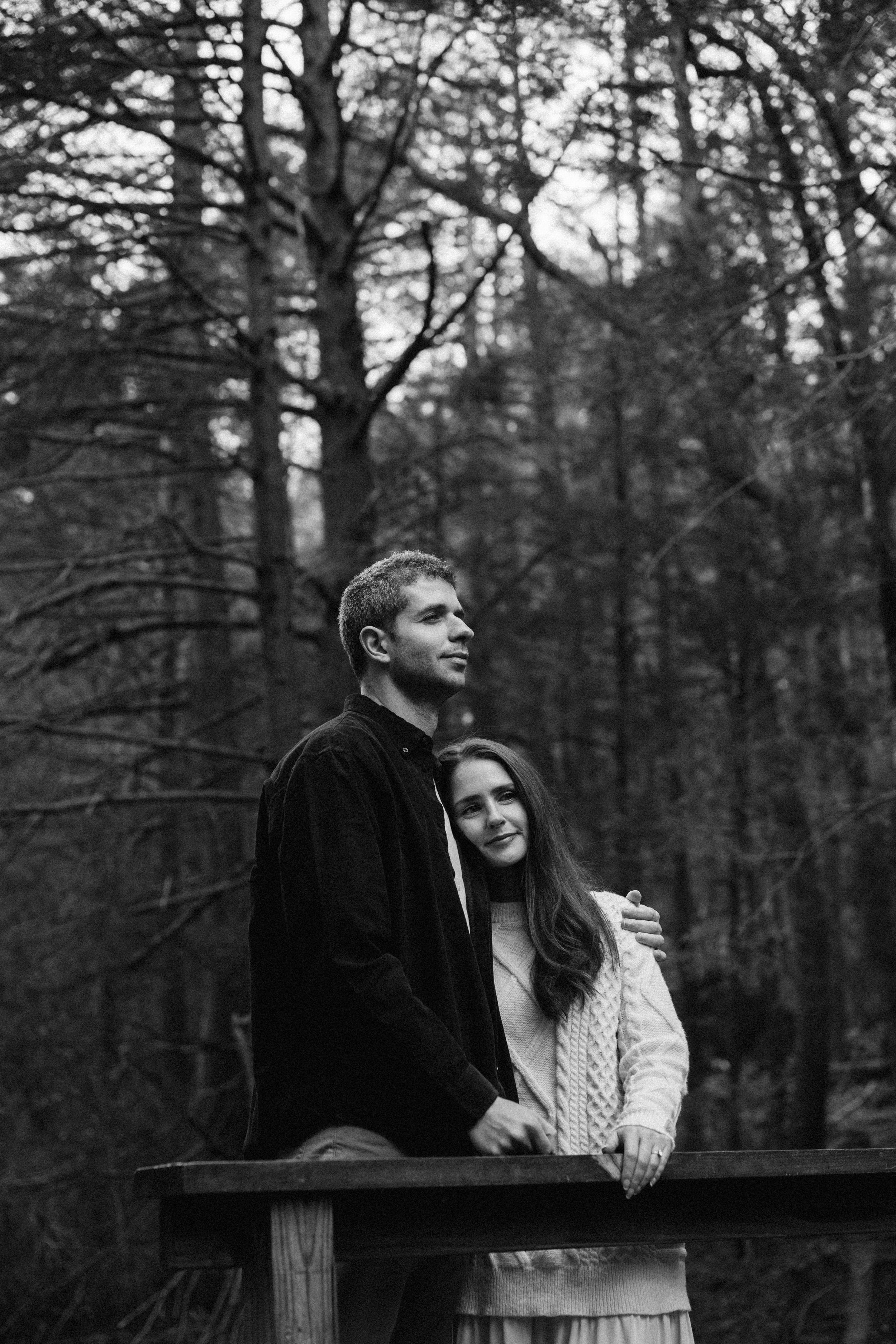 Black and white photo of a couple embracing on a wooden bridge surrounded by bare trees in a forest.