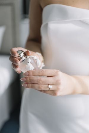 Bride in white strapless gown holding perfume bottle with tassel, wearing solitaire diamond engagement ring.