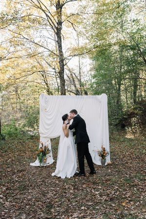 Bride and groom share first kiss at outdoor fall wedding ceremony with white draped backdrop and autumn floral arrangements.