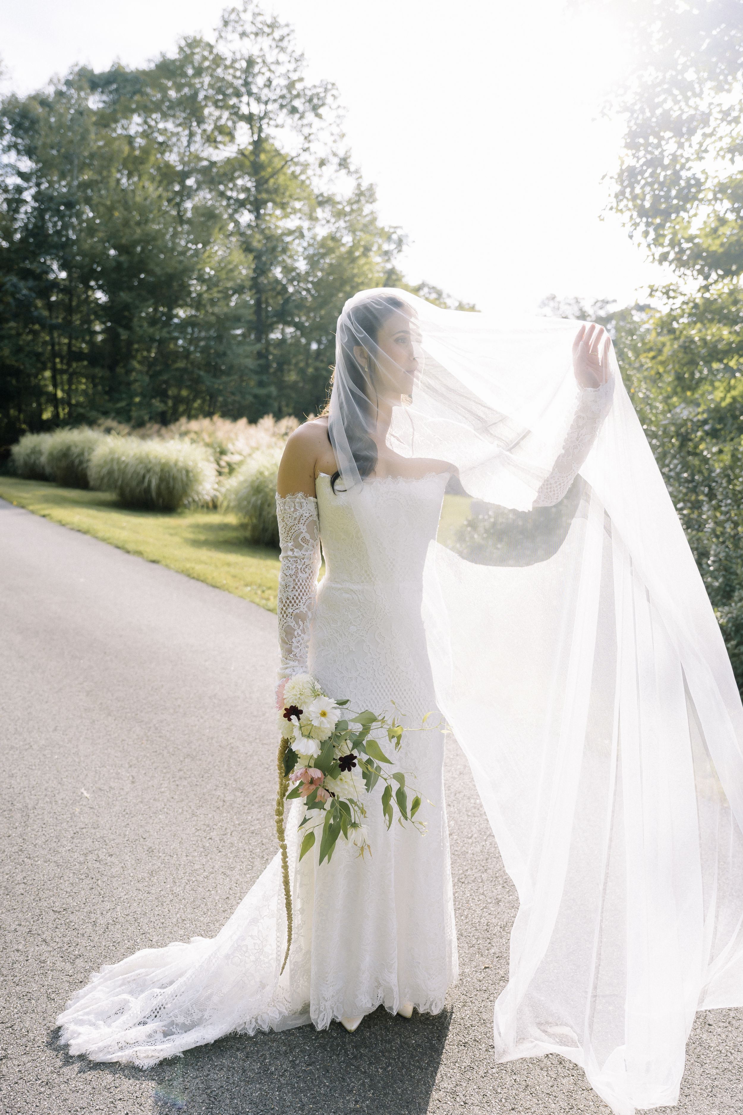 Bride in white lace gown and veil holds bouquet on sunlit path surrounded by lush greenery.
