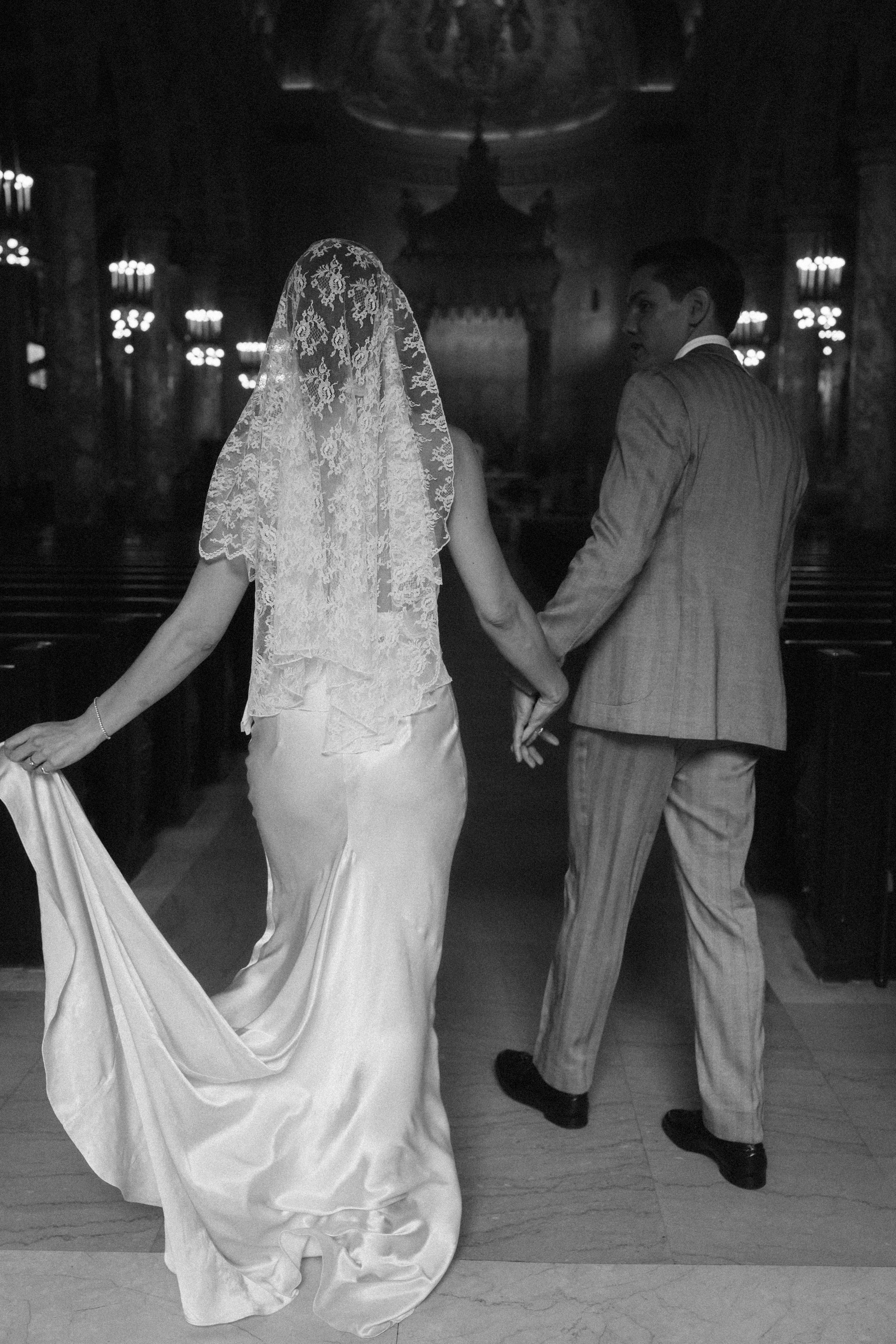 Black and white photo of bride and groom holding hands walking down church aisle in elegant wedding attire.