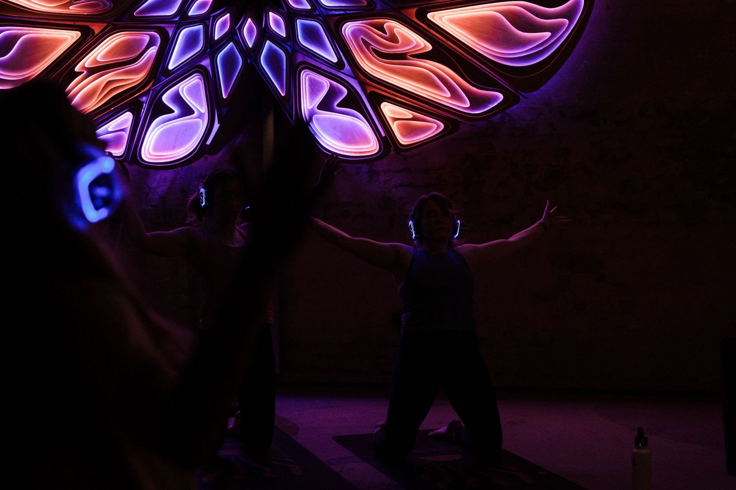 Silhouettes of people dancing under vibrant neon purple and orange glowing mandala art installation in dark room.