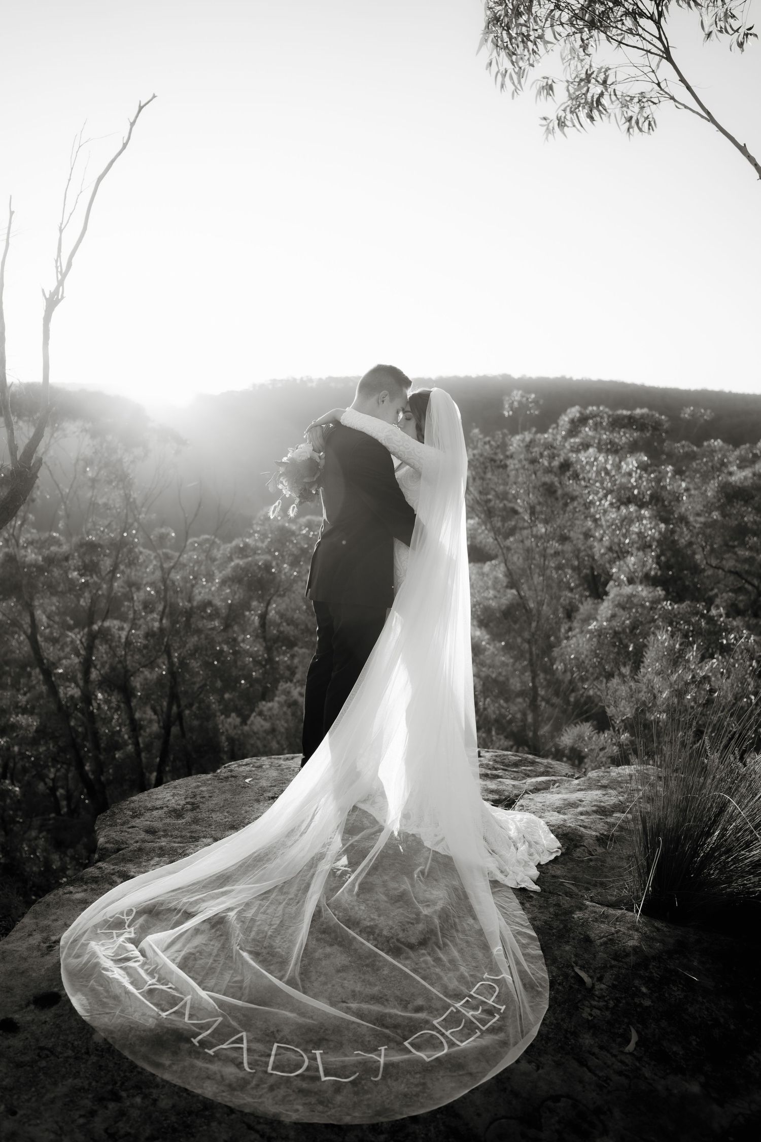 Black and white wedding photo of bride and groom embracing on rocky cliff, long veil trailing with text, surrounded by bushland.