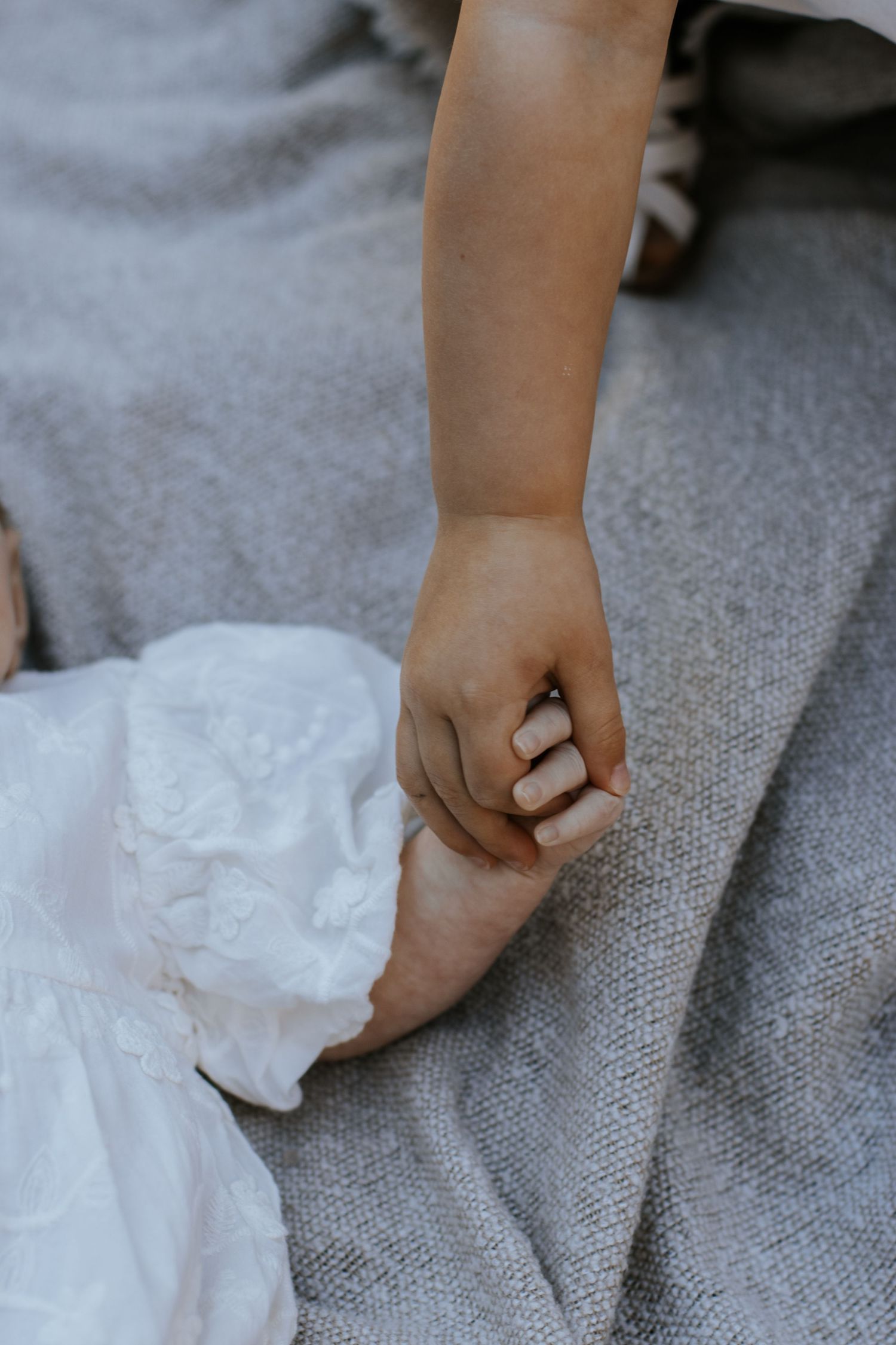 A small child's hand gently gripping an adult's finger, wearing a white dress on a gray blanket.