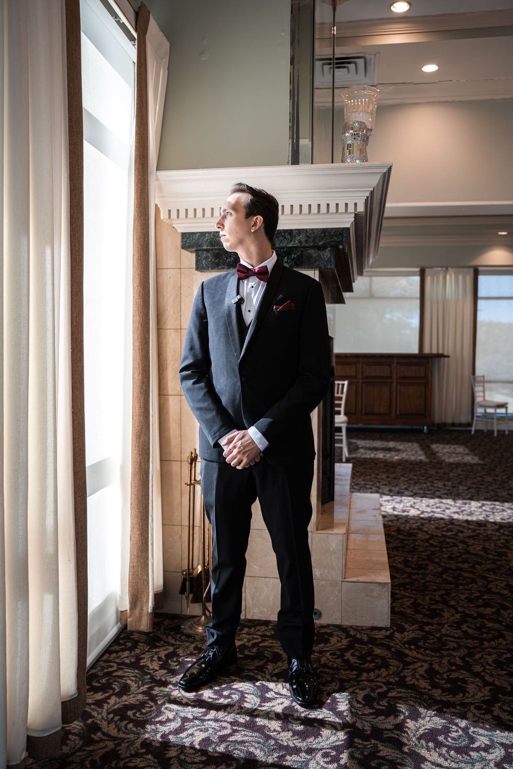 Groom in black tuxedo with burgundy bow tie stands by window in elegant venue with fireplace.