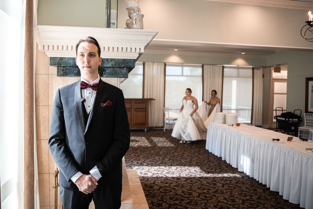 Groom in gray suit with burgundy bow tie stands in elegant ballroom as bride in white gown walks in background.
