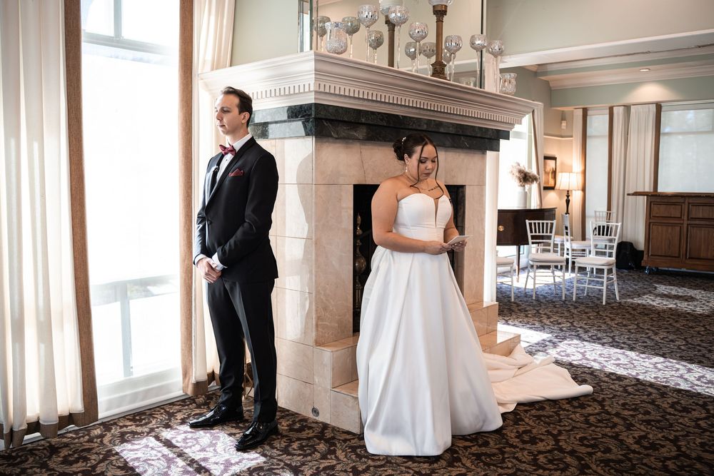 Bride and groom stand back-to-back in elegant venue with fireplace during wedding first look moment.