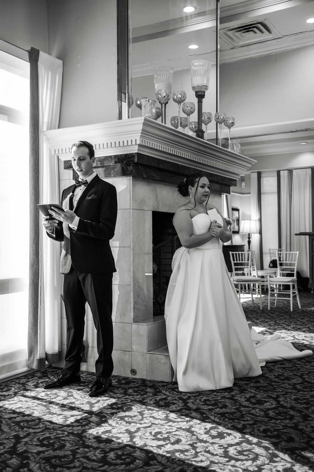 Black and white photo of bride and groom reading vows separately near elegant fireplace in wedding venue.