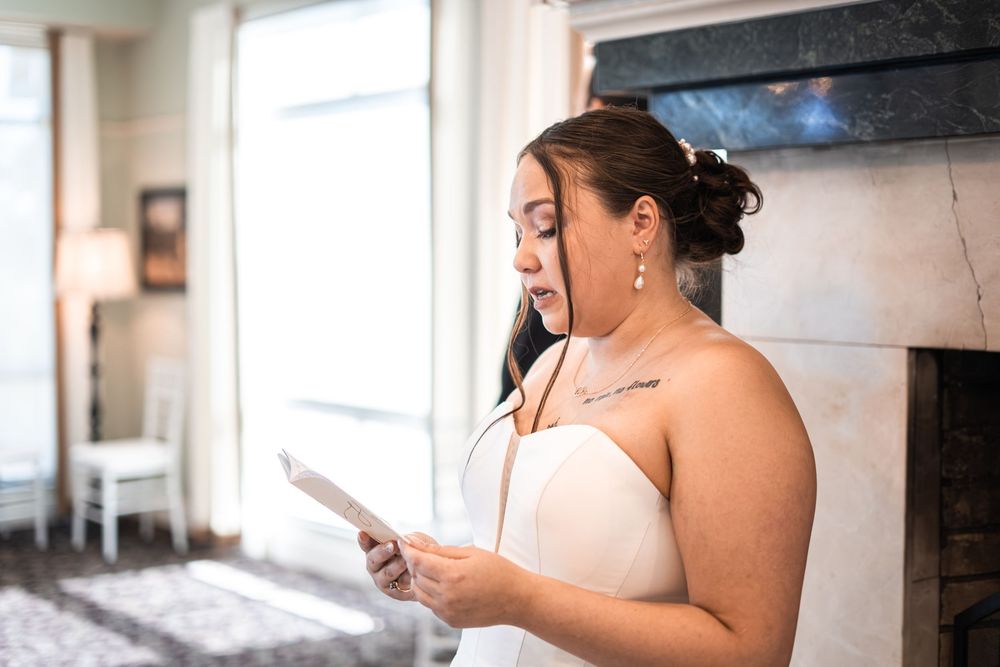 Bride in strapless white gown reads wedding vows from paper near fireplace during bridal preparation.