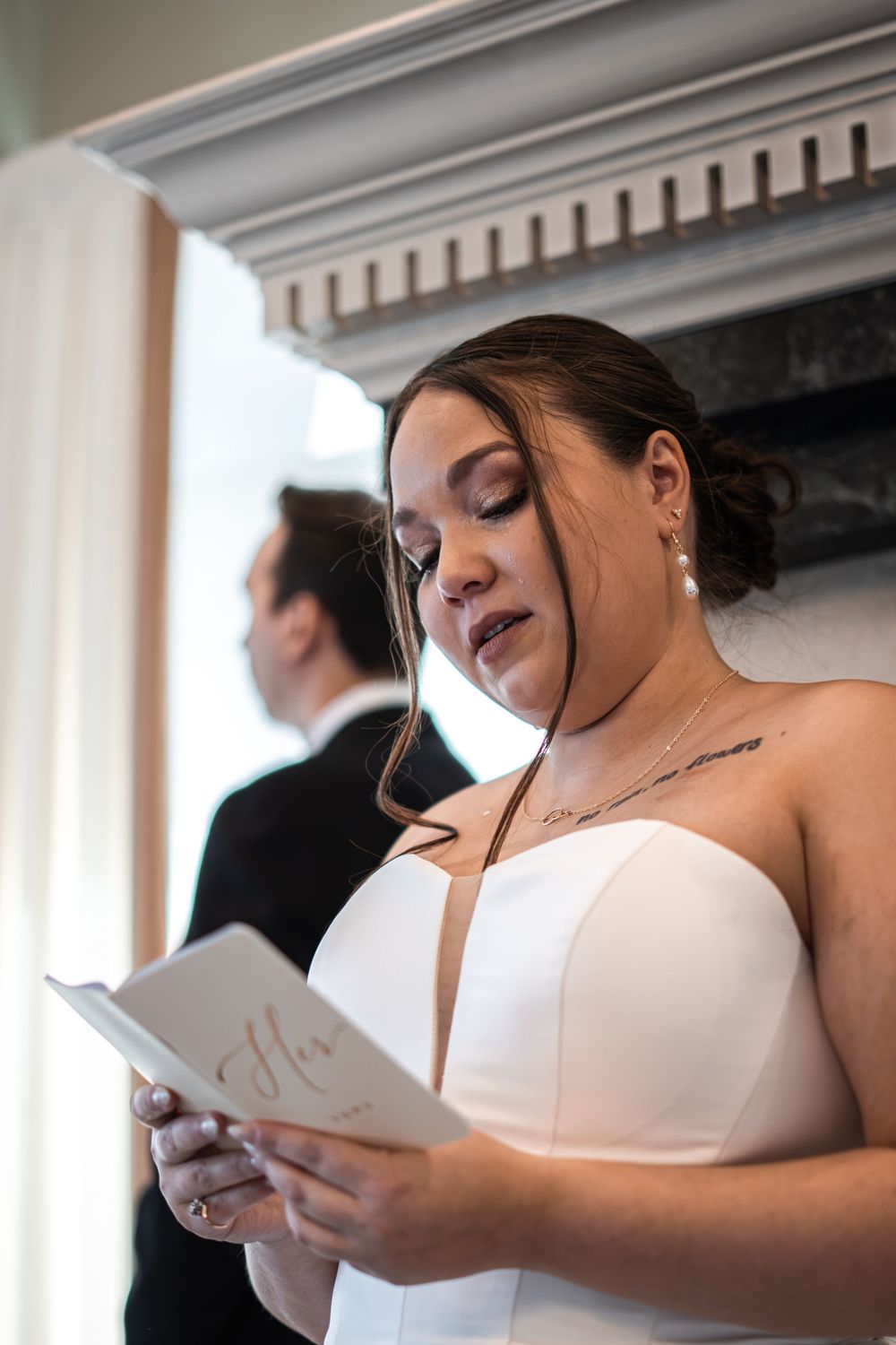 Bride in strapless white gown reads wedding vows from a card, with groom visible in background near fireplace.