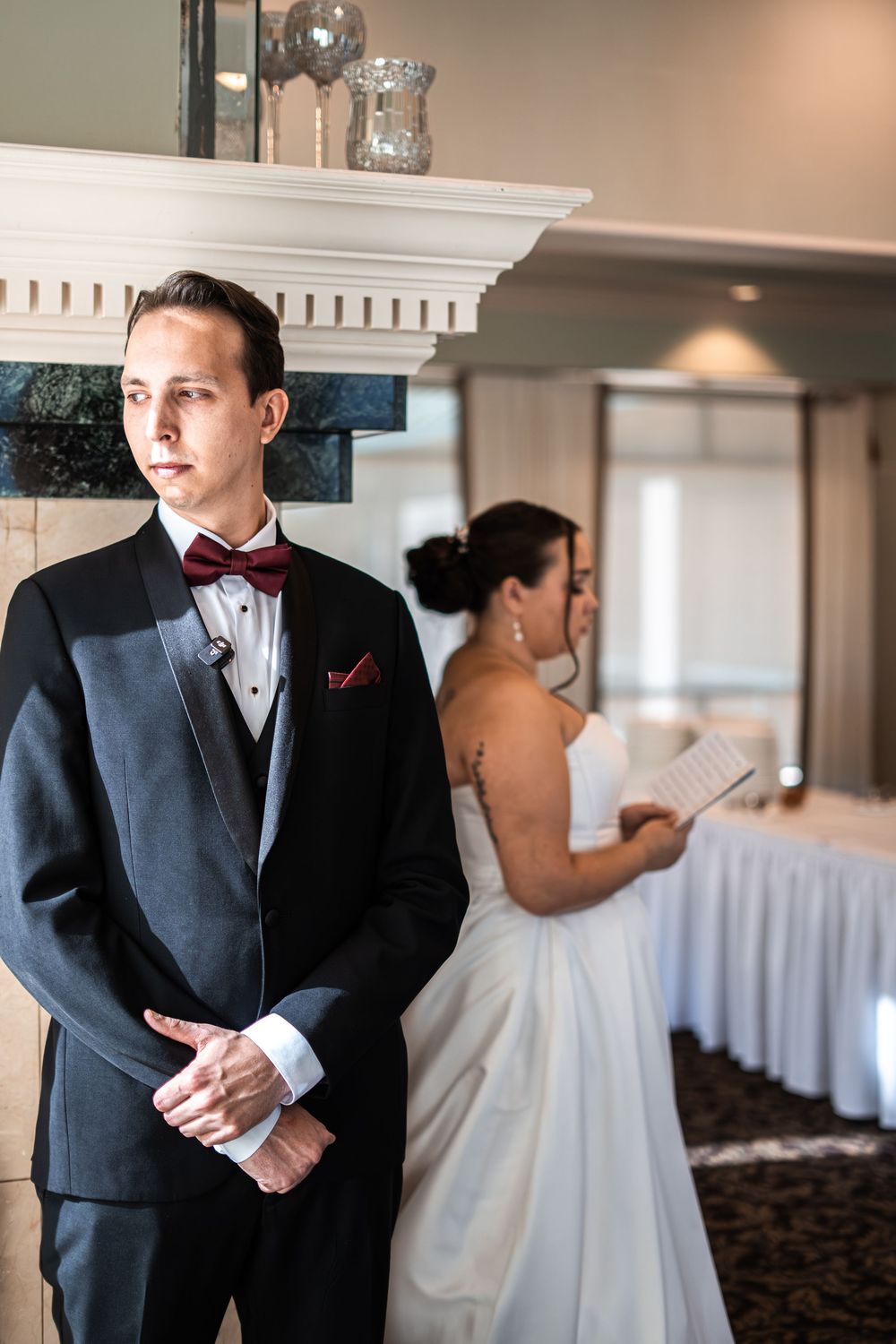 Groom in black tuxedo with burgundy bow tie stands before fireplace as bride in white gown reads vows behind him.