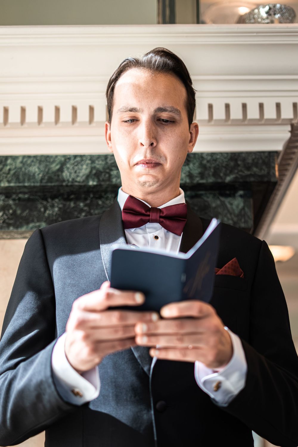 Emotional groom in black tuxedo reads wedding vows from small booklet near elegant marble fireplace.