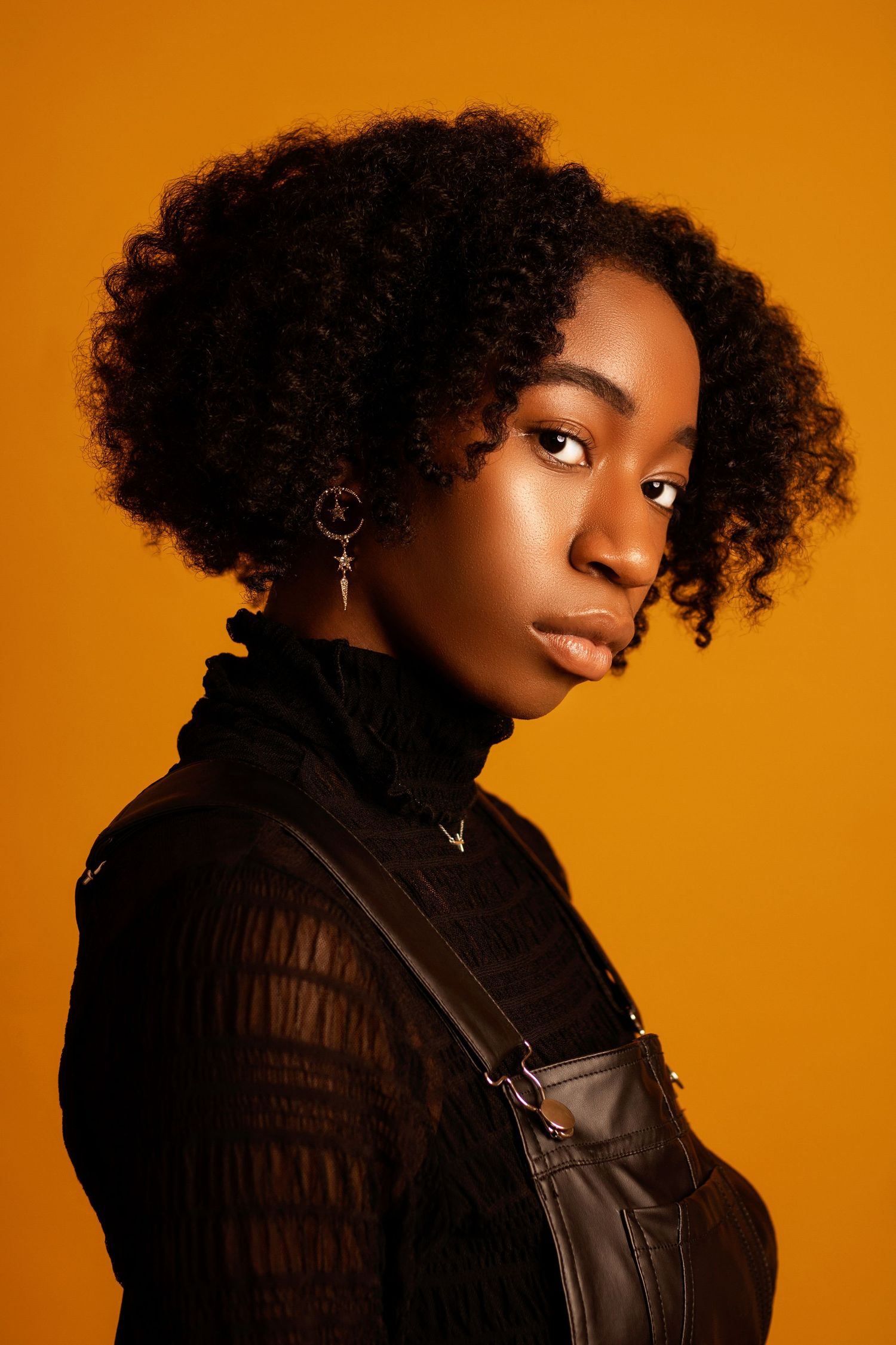 Young Black woman with curly afro hair wearing black turtleneck and overalls, posing against vibrant orange background.