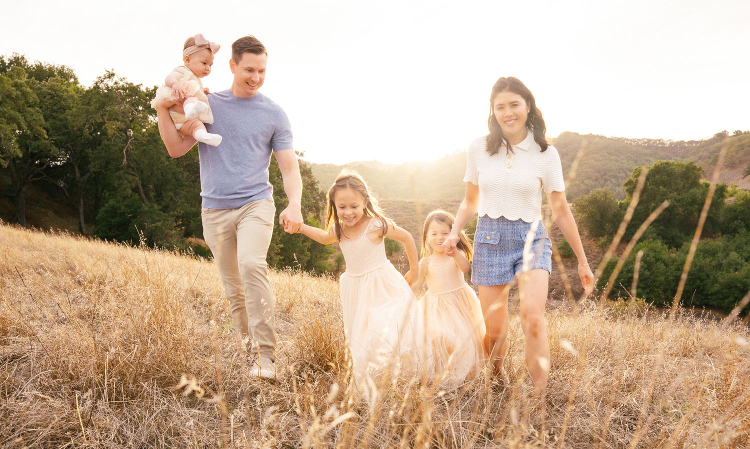 Happy family of five walking through golden field at sunset, parents holding hands with young daughters in pink dresses.