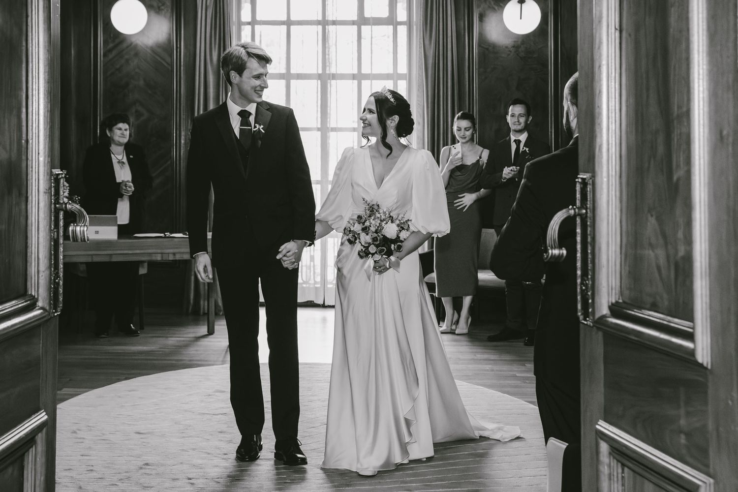 Black and white photo of a bride and groom holding hands walking down the aisle at an elegant wedding ceremony.