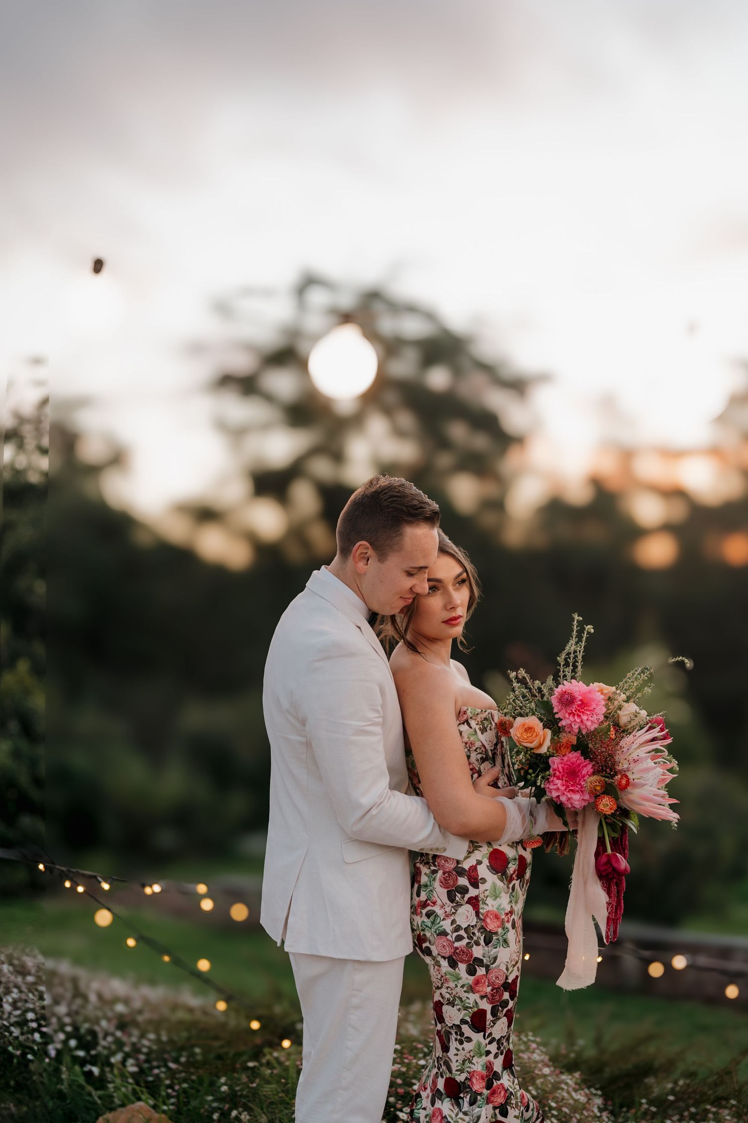 Couple embracing at sunset, bride in floral dress holding vibrant bouquet, groom in white suit, string lights glowing in background.