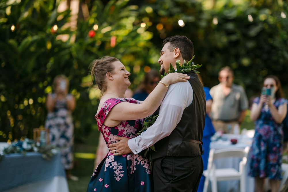 Couple shares first dance outdoors at garden wedding, groom wearing lei, guests watching in background.