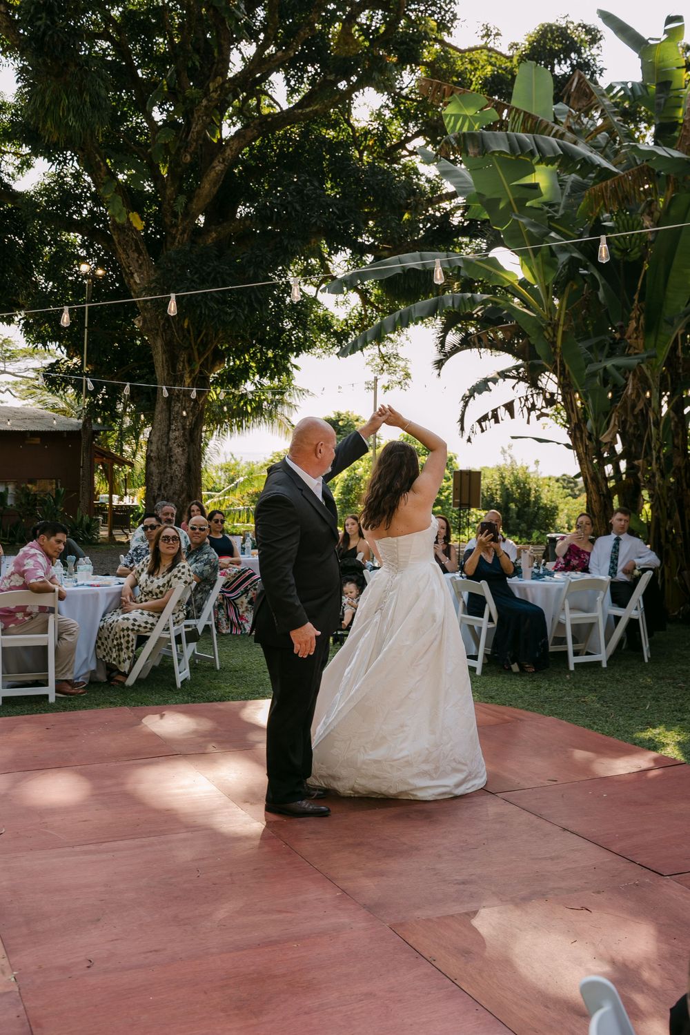Bride and groom share first dance on outdoor wooden floor surrounded by guests at a garden wedding reception.