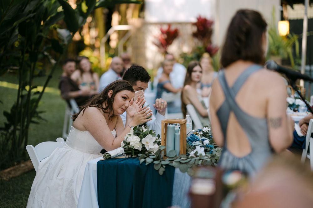 Bride wipes tears at decorated reception table while guests listen to wedding speech outdoors.