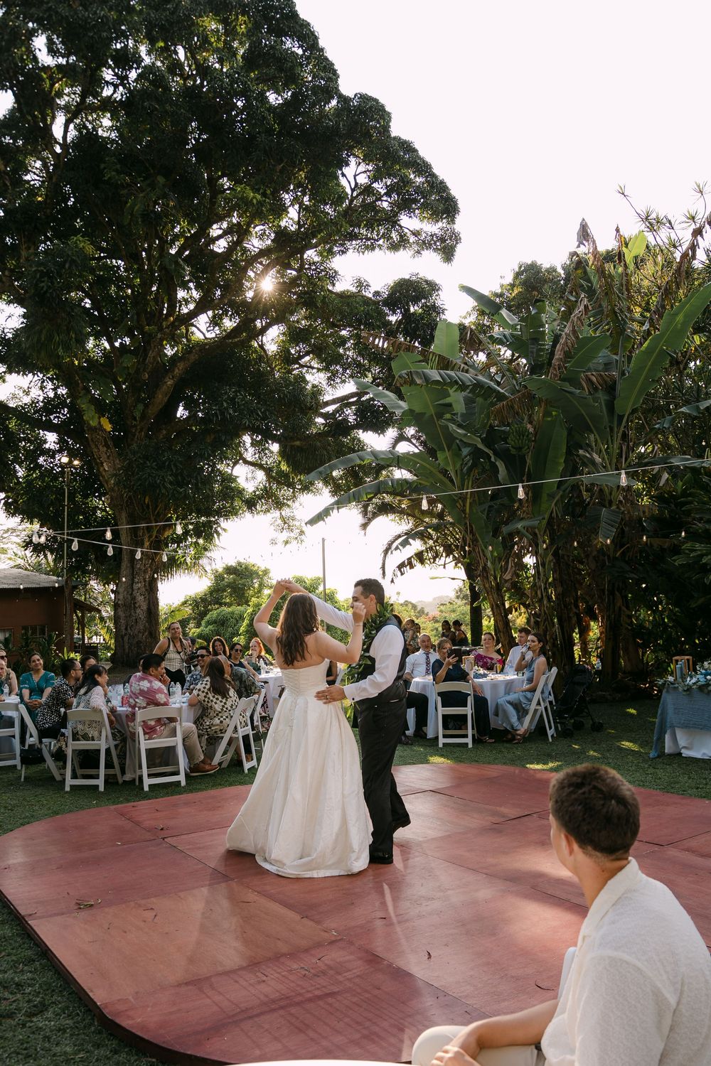 Bride and groom share their first dance on an outdoor wooden floor surrounded by guests at a garden wedding reception.