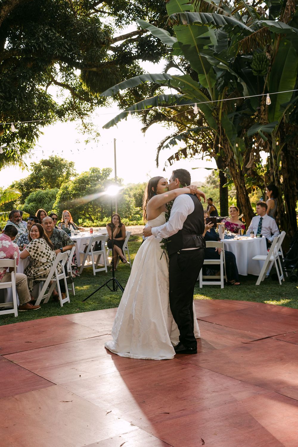 Bride and groom share first dance kiss on outdoor red dance floor surrounded by guests at a garden wedding reception.