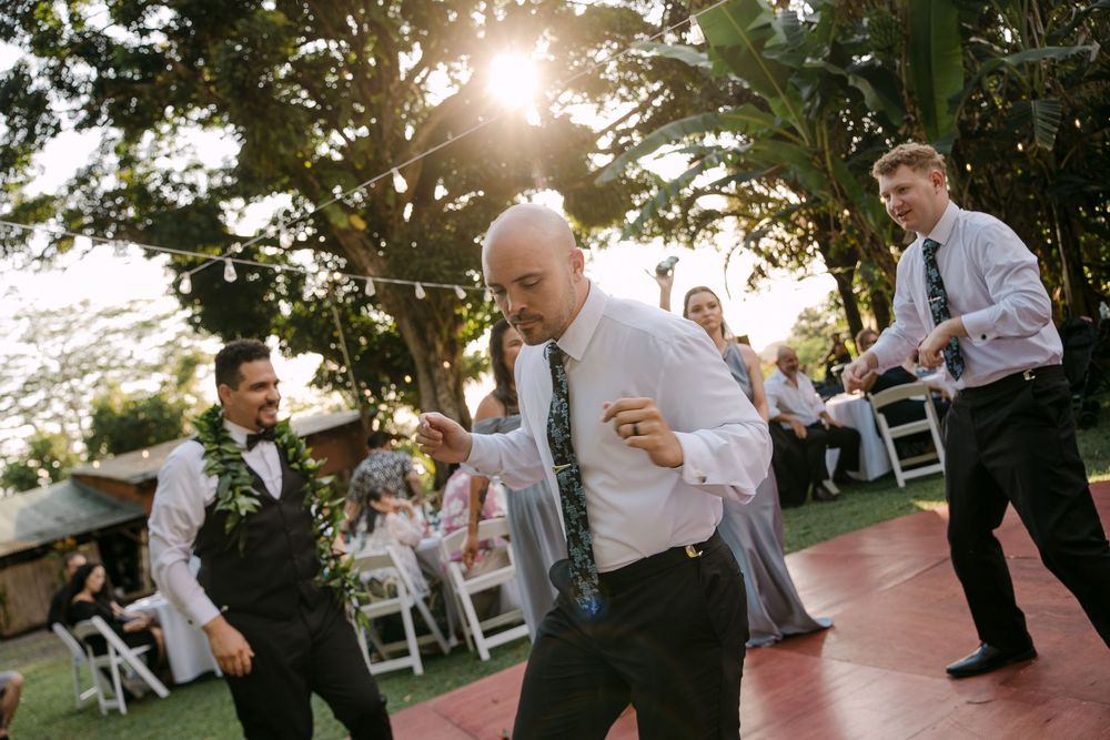 Men dancing at an outdoor wedding reception on a red dance floor surrounded by lush green trees.