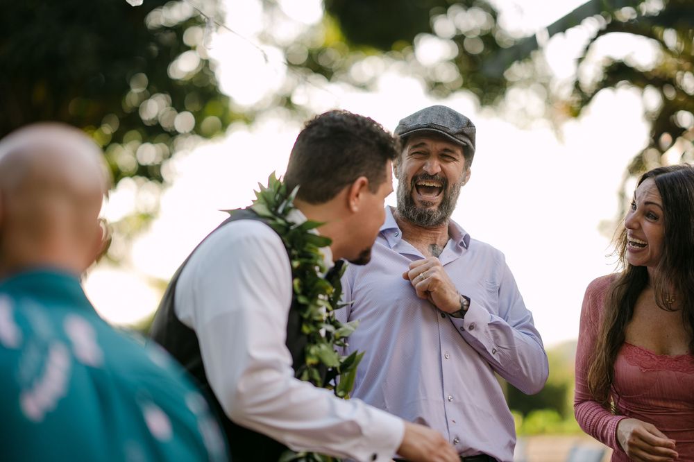 Guests laughing and celebrating at an outdoor wedding reception, with a groom wearing a floral lei.