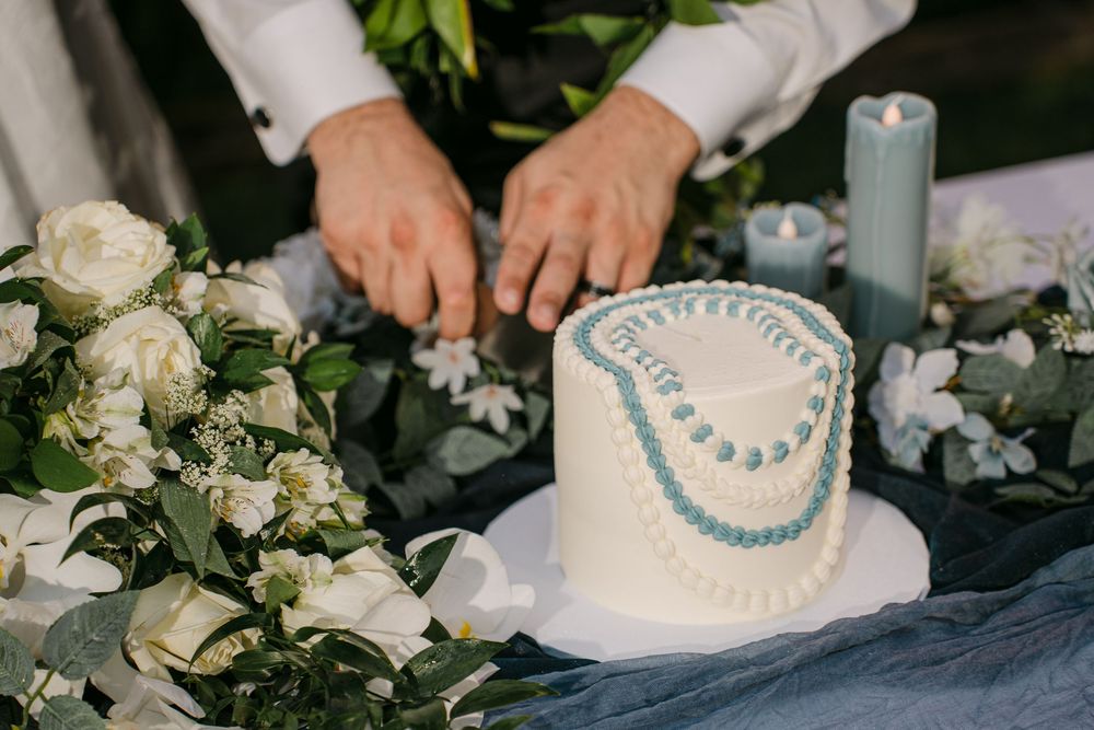 Hands cutting a heart-shaped white wedding cake with blue piping, surrounded by white flowers and candles.