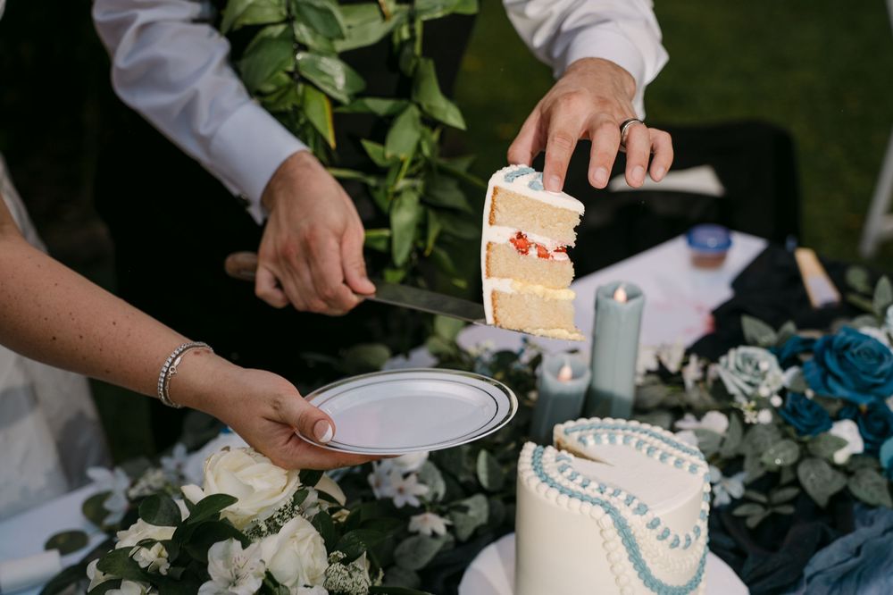 Bride and groom cutting a white wedding cake with blue floral decorations at an outdoor reception.