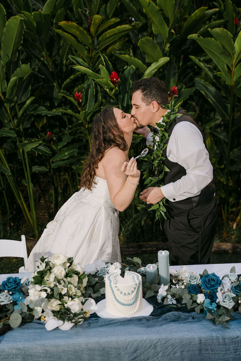 Bride and groom sharing a kiss while cutting their wedding cake outdoors, surrounded by tropical greenery and blue floral arrangements.