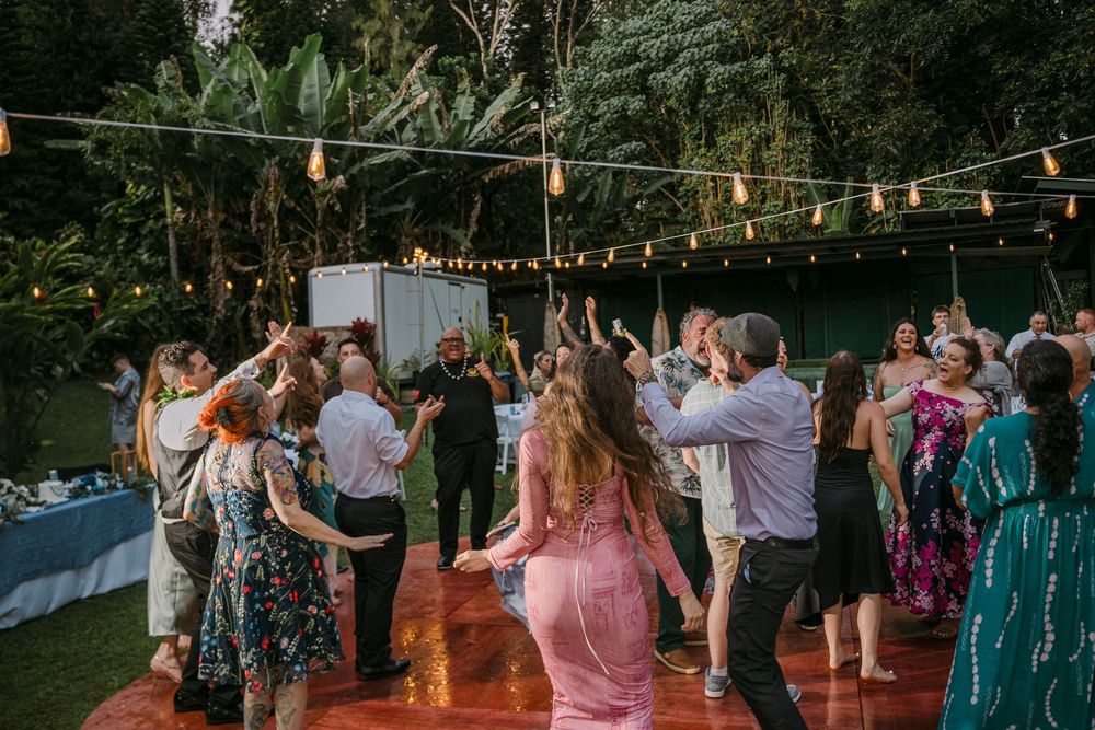 Guests dancing outdoors at a festive evening wedding reception with string lights and lush greenery backdrop.