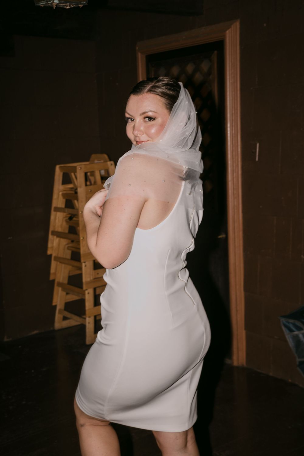 Bride in white mini dress and veil poses elegantly in dimly lit venue, glancing over shoulder.