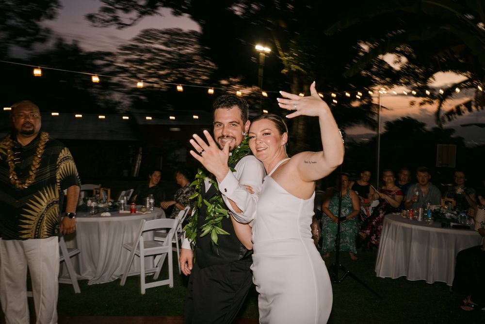 Joyful newlyweds wave to guests during outdoor wedding reception at dusk with string lights.