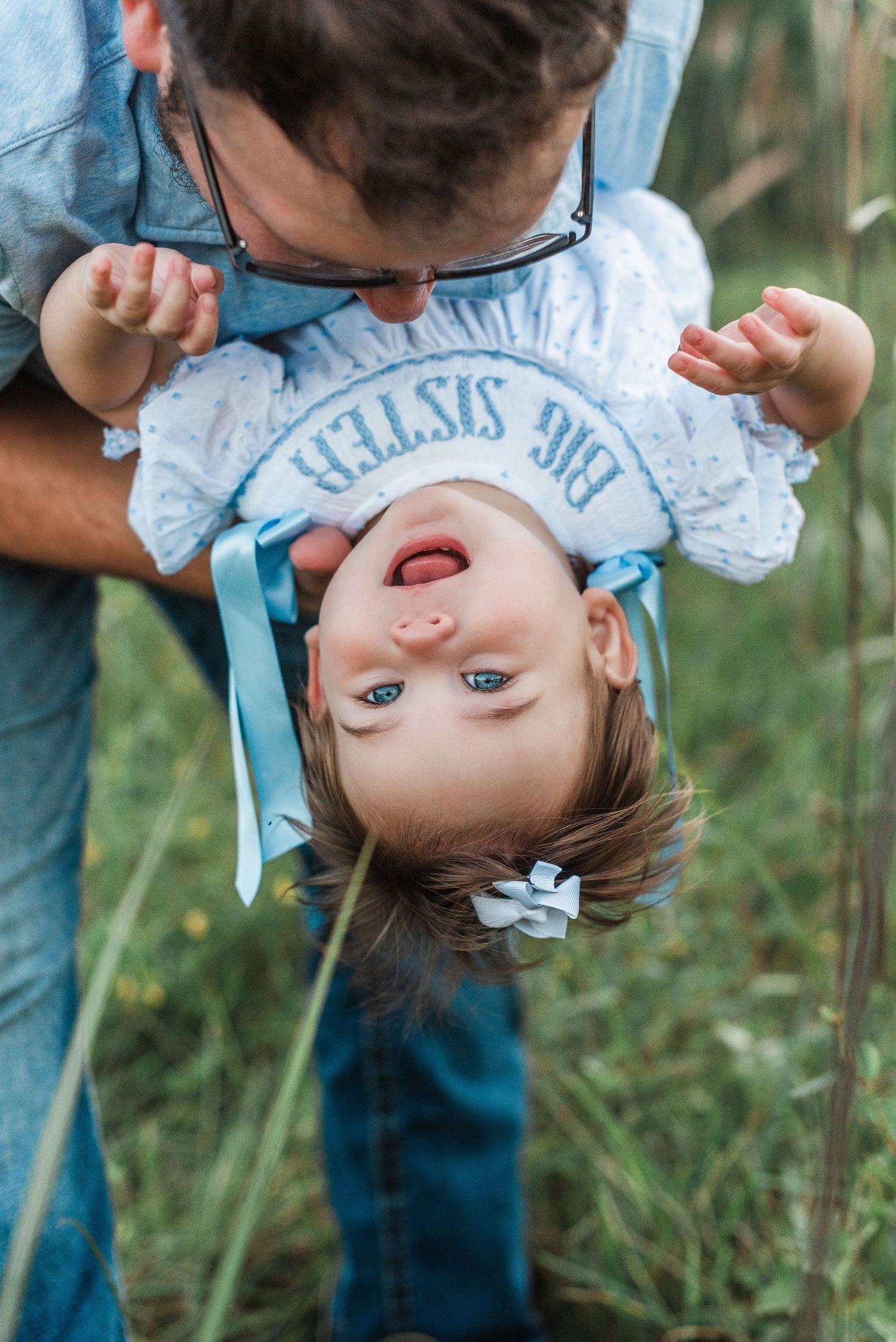 A playful upside down shot of a small child and her dad in blue and white clothing against a natural outdoor background.