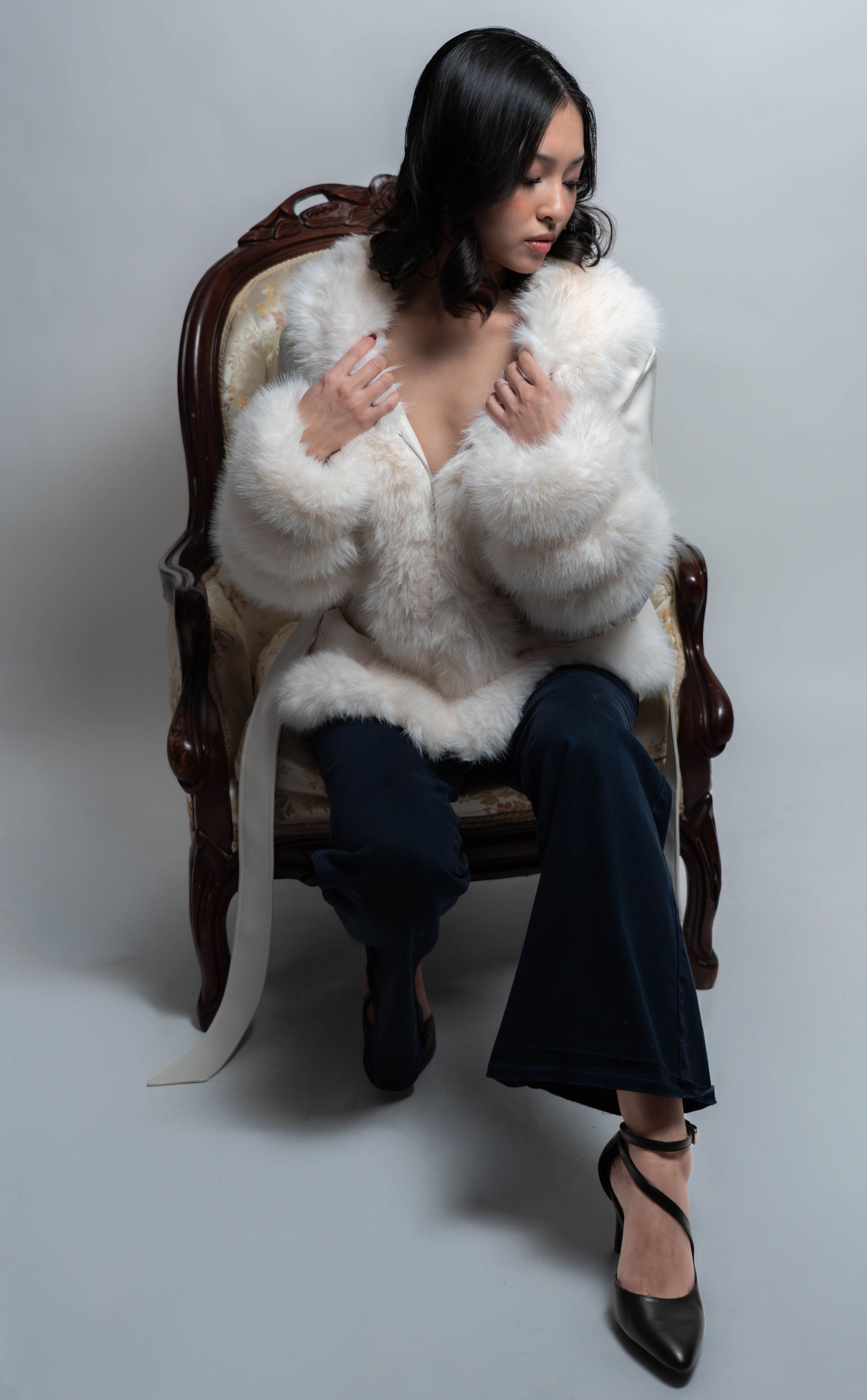 Woman in white fur coat and dark flare jeans seated on ornate chair, wearing black heeled shoes.