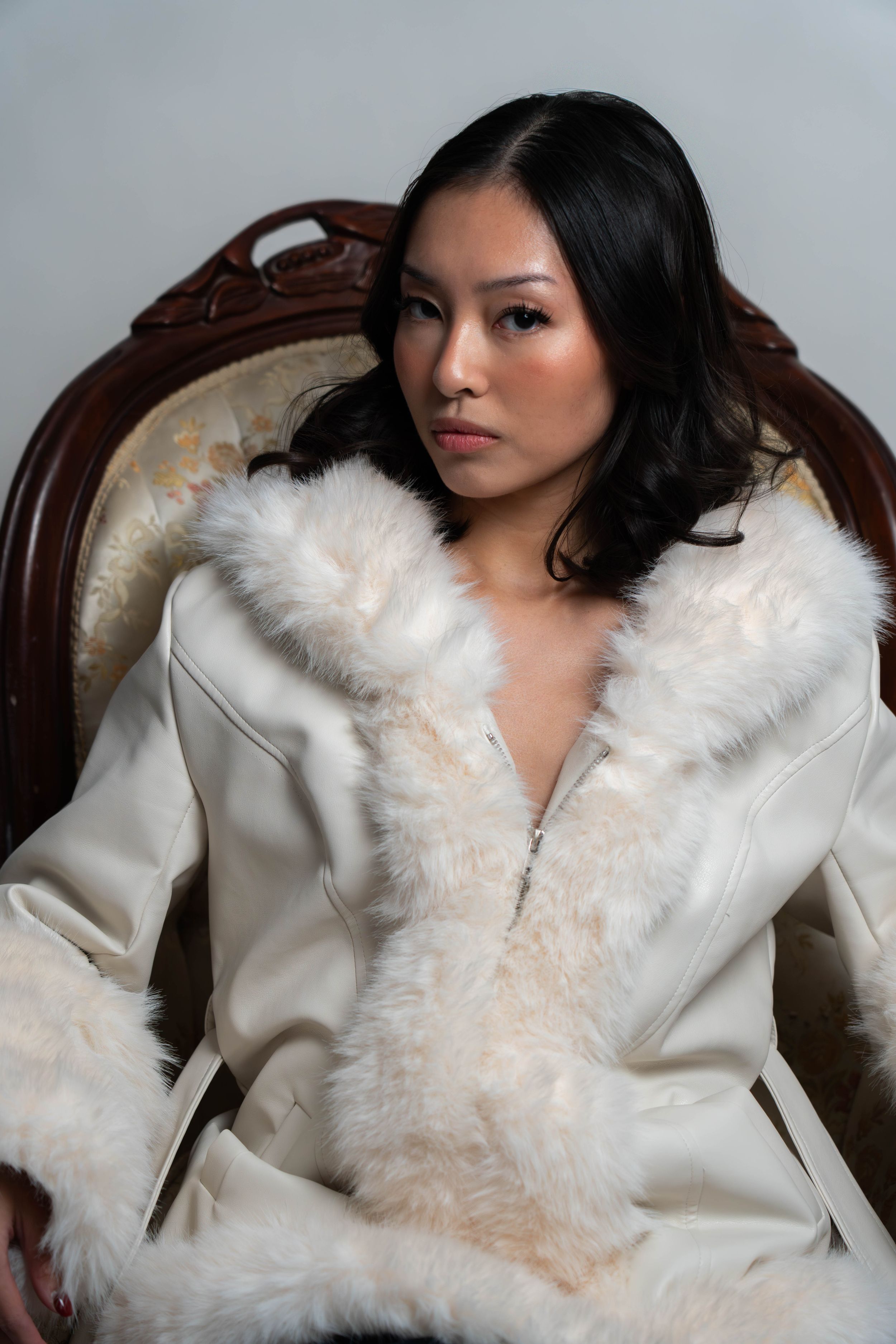 Woman in white fur-trimmed coat seated in ornate vintage chair, posing elegantly against gray background.
