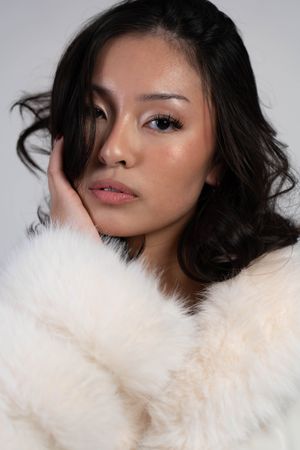Young woman with dark wavy hair poses in a luxurious white fur coat, resting her hand gently against her cheek.