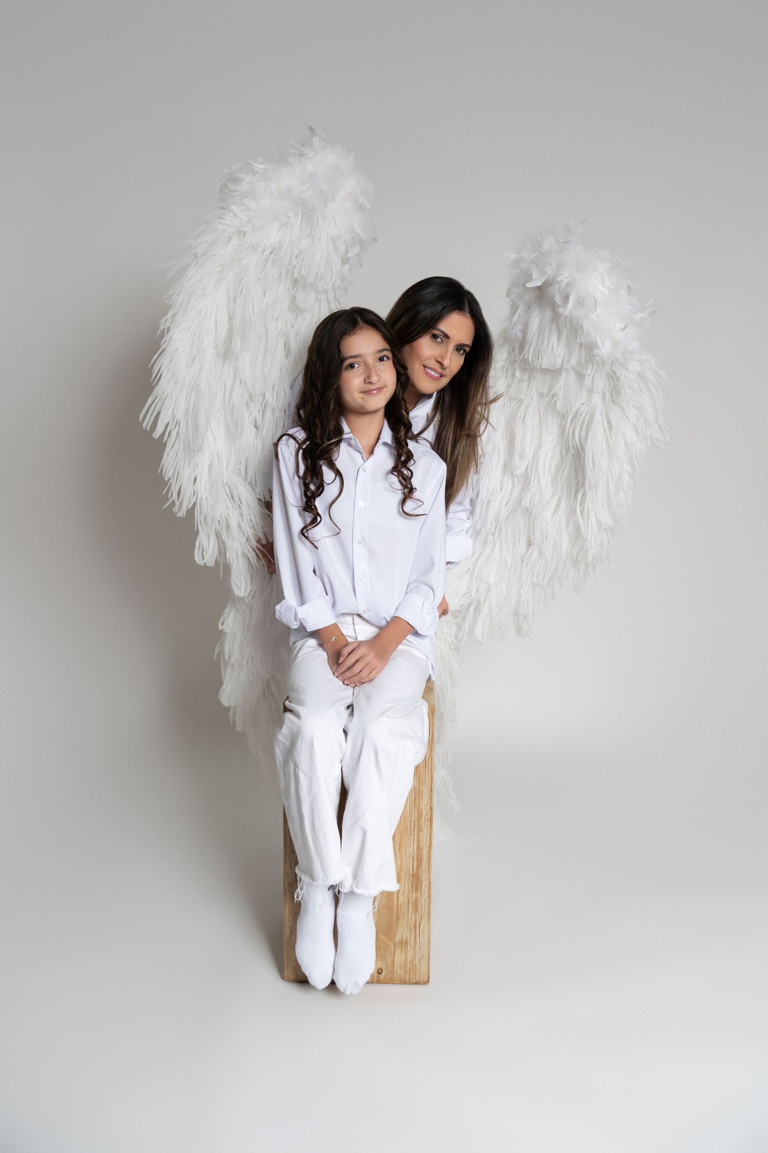 Two women in matching white shirts and pants sit back-to-back on a studio floor, smiling warmly.