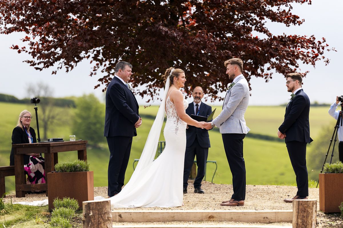Couple exchanging vows at an outdoor wedding ceremony with guests seated on either side in a scenic countryside setting.