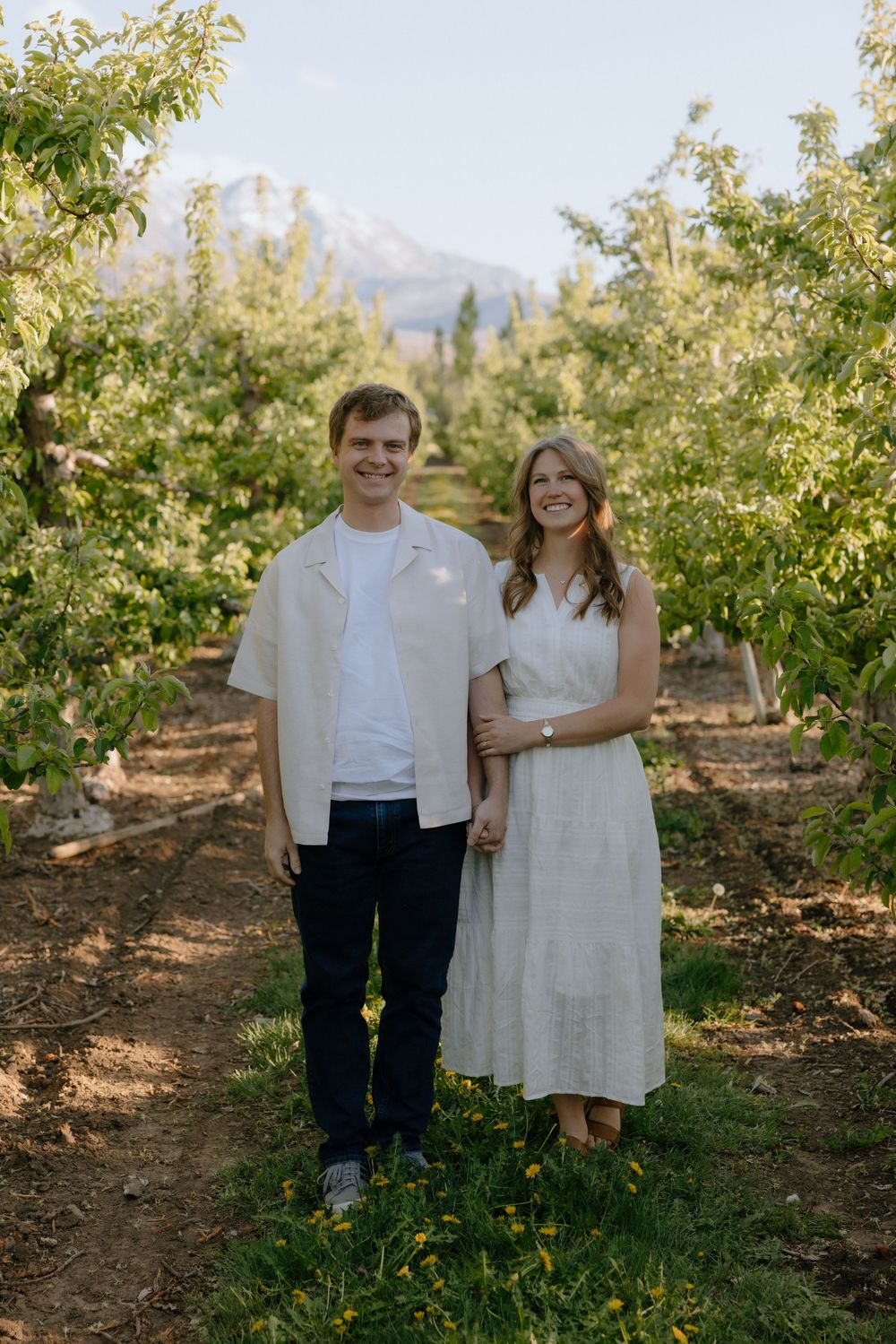 Couple smiling together in a lush orchard with a mountain backdrop on a sunny day.