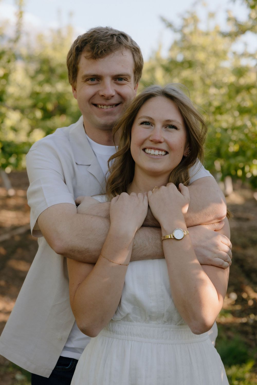 Smiling couple embracing in a lush green orchard, woman in white dress, man in beige jacket.