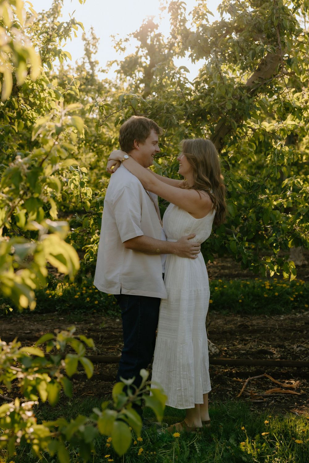 Couple embracing and smiling at each other in a sunlit green orchard, woman in white dress, man in white shirt.