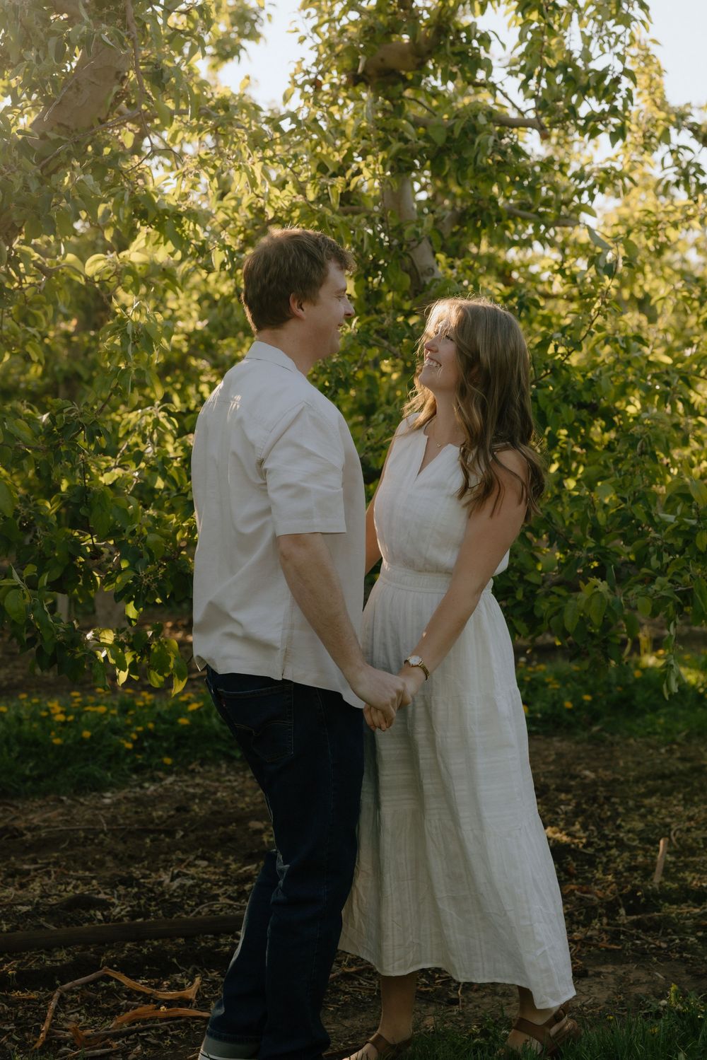 Couple holding hands and smiling at each other in a sunlit orchard, wearing white outfits.