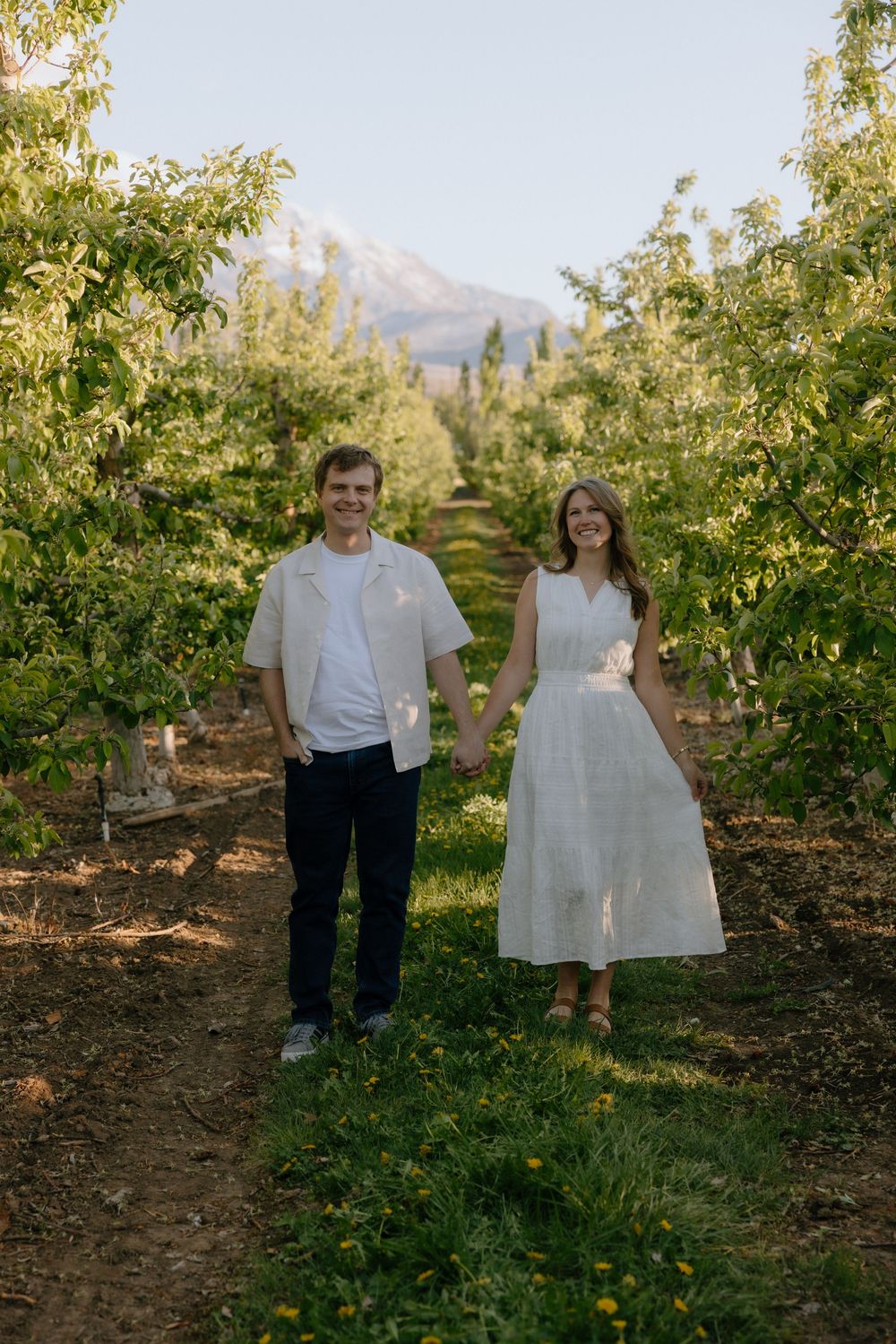 Couple holding hands in a sunlit orchard with mountain backdrop, wearing white outfits.