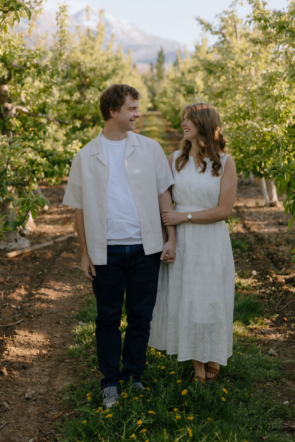 Couple holding hands and smiling at each other in a lush green orchard with mountains in the background.