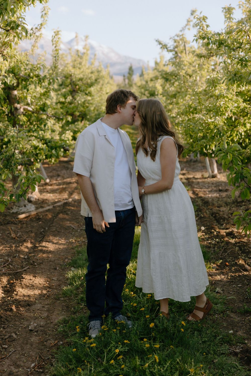Romantic couple sharing a kiss in a green orchard with a mountain visible behind them.