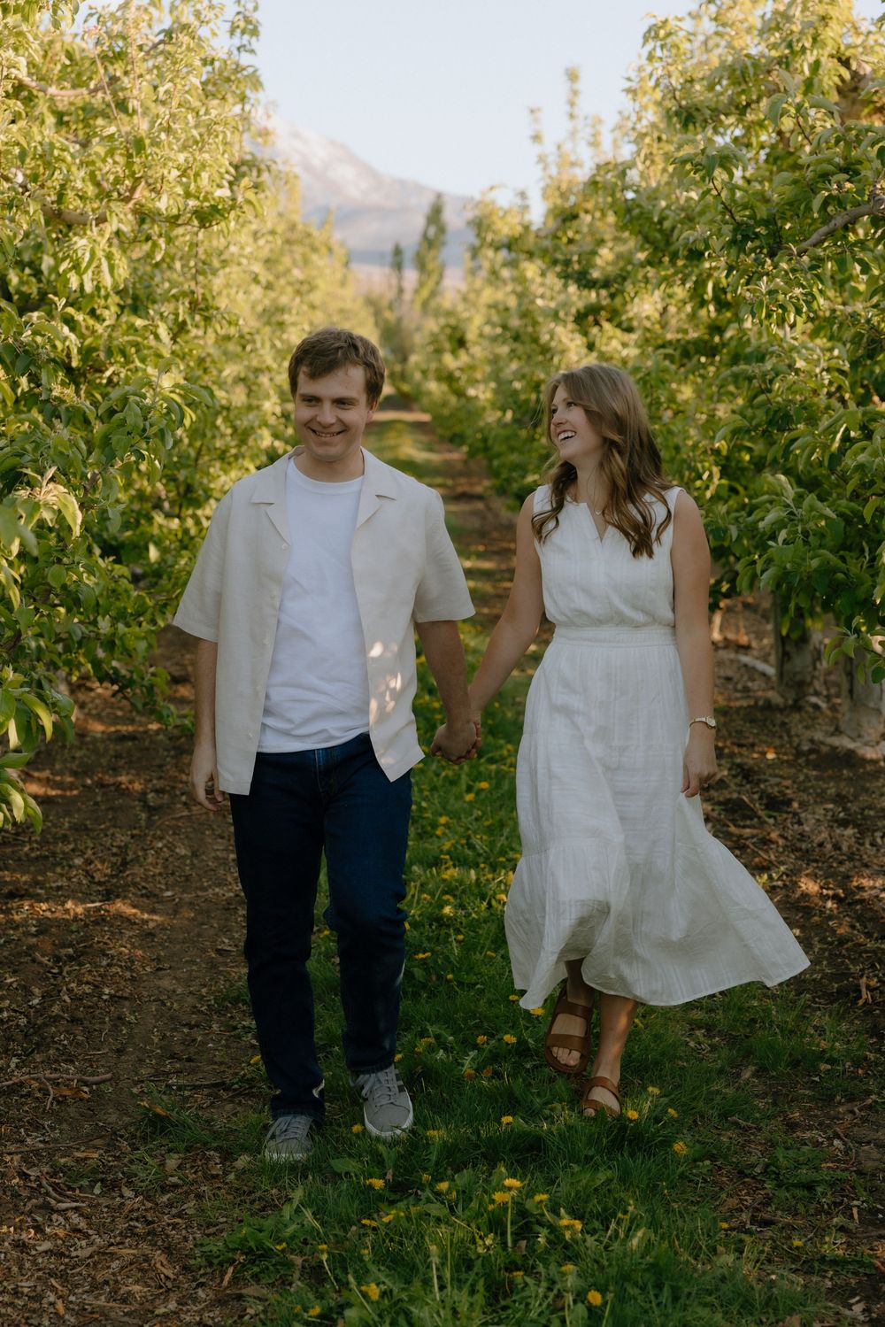 Happy couple walking hand in hand through a lush green orchard at golden hour.