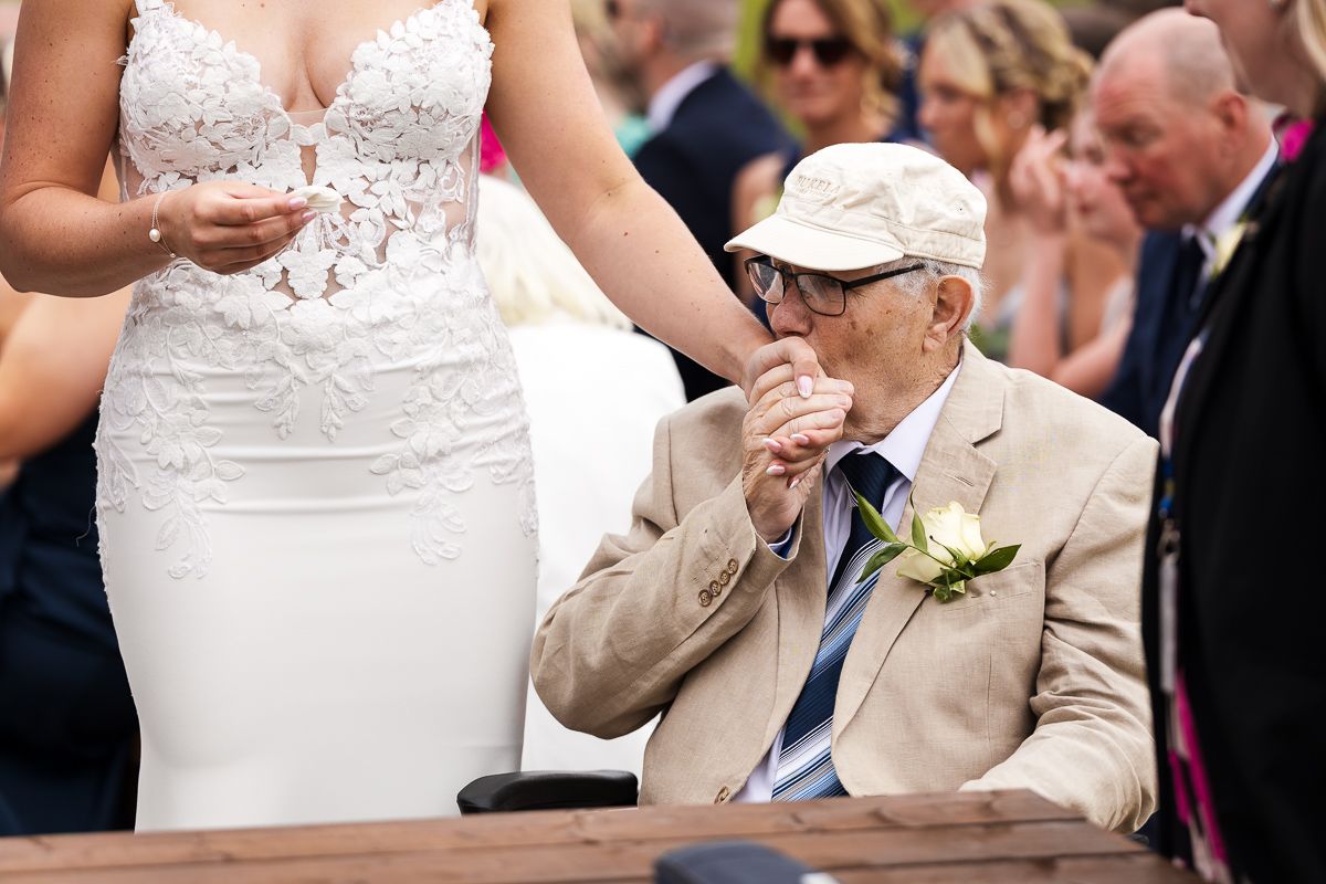 Emotional wedding guest in beige suit and cap wipes tears during ceremony while seated at pew.