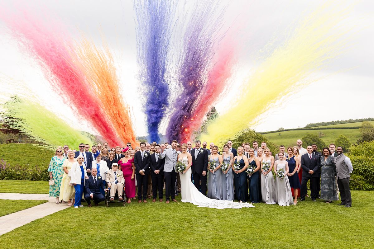 Wedding group photo with colorful rainbow smoke bombs exploding above guests on a green lawn.